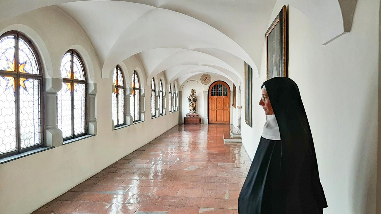 Das Bild zeigt einen schmalen, hellen Flur im Benediktinerinnenkloster Nonnberg. Die Wände des Flurs sind in einem neutralen, hellen Farbton gehalten. An der linken Seite sind mehrere große, gotische Fenster mit bunten Glasmalereien, die jeweils einen goldenen Stern zeigen. Der Boden besteht aus glänzenden, terrakottafarbenen Fliesen, die gut gepflegt wirken.  Am Ende des Flurs steht eine Holzstatue einer heiligen Figur auf einem Podest, rechts daneben befindet sich eine Holztür, die ins nächste Zimmer führt. Auf der rechten Seite des Flurs steht eine Frau in traditioneller Klosterkleidung, die eine schwarze Robe und eine weiße Haube trägt. Sie schaut nachdenklich in die ferne und ist von Profil zu sehen. Die Beleuchtung des Flurs ist freundlich und weich, was eine ruhige Atmosphäre schafft.