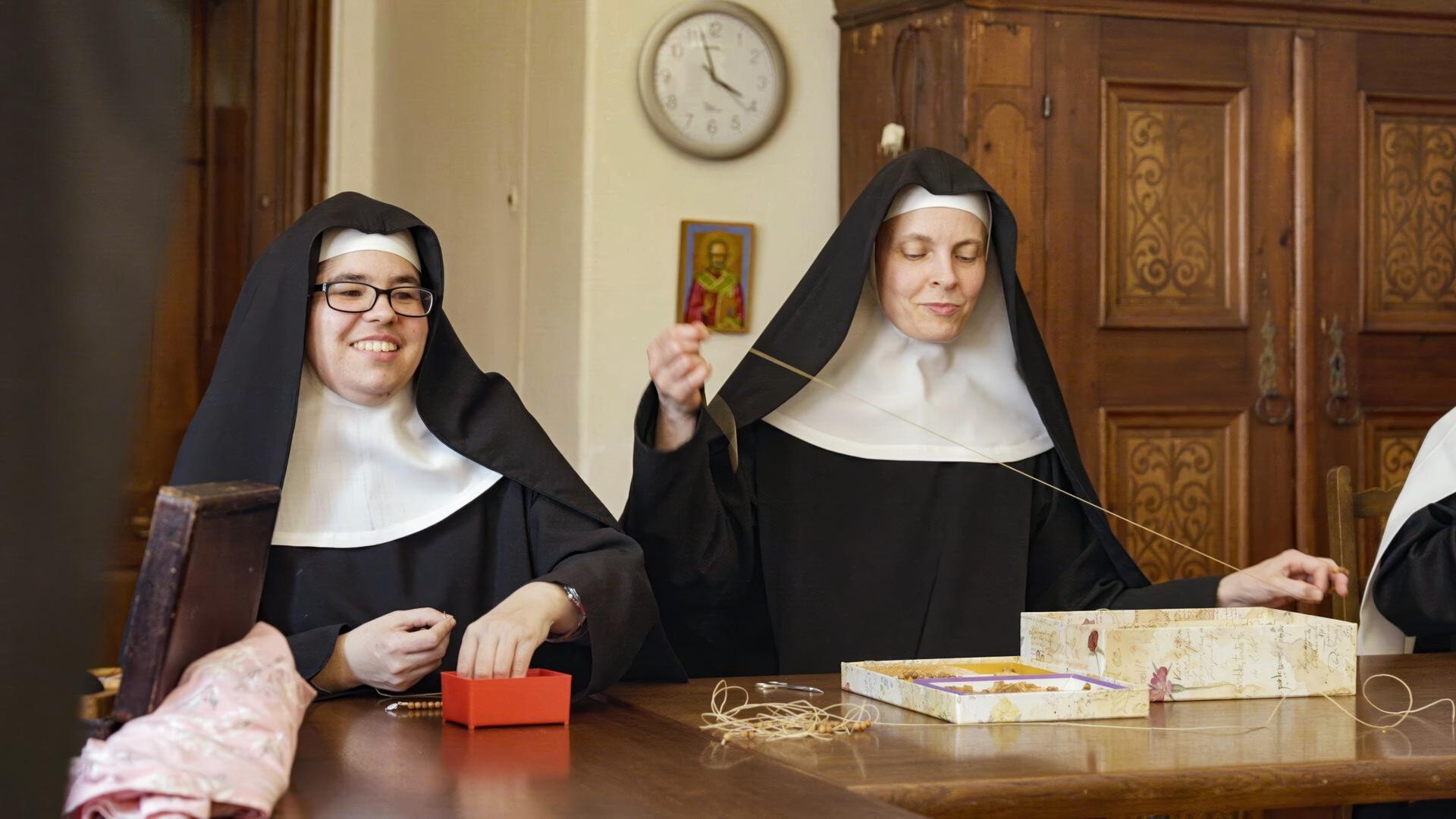 Das Bild zeigt zwei klosterbekleidete Frauen, die in einem Raum sitzen, der ein warmes, einladendes Ambiente ausstrahlt. Beide tragen traditionelle Habitbekleidung, bestehend aus schwarzen Gewändern und weißen Hauben. Die Frau auf der linken Seite hat eine Brille und lächelt, während sie an einem kleinen roten Kästchen sitzt. Ihre Hände sind beschäftigt, wahrscheinlich mit einer Art Bastelarbeit oder Hobby. Die andere Frau, die sich rechts befindet, hält einen Faden in der Hand und sieht konzentriert aus.