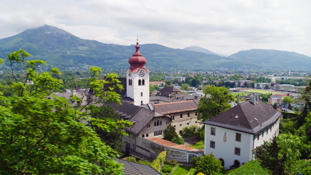 Das Bild zeigt die Benediktinerinnenabtei Nonnberg in Salzburg. Im Vordergrund ist ein Teil des Klosters zu sehen, das aus mehreren Gebäuden mit dunklen Dächern und hellen Fassaden besteht. Besonders auffällig ist der Kirchturm mit einer roten Kuppel und einem Ziffernblatt. Um das Kloster herum befinden sich Bäume, die das Bild rahmen. Im Hintergrund erheben sich sanfte, bewaldete Hügel und Berge, während der Himmel leicht bewölkt ist.  Die Abtei Nonnberg ist das älteste Frauenkloster im deutschsprachigen Raum und liegt am Fuß der Festung Hohensalzburg. Die Landschaft rund um das Kloster ist grün und zeigt Anzeichen von ländlicher Bebauung.