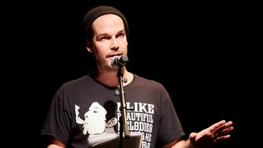 Ein junger Mann mit Mütze steht vor einem Mikrofon und trägt etwas aus einem Buch vor.