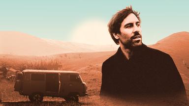 Ein Porträt von Max Giesinger vor der Wüste, neben ihm ist ein Camper zu sehen.