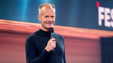 Max Uthoff steht auf der linken Seite des Bildes. Er hält ein Mikrofon in seiner rechten Hand. Die Bühne hinter ihm ist unten rot und oben blau beleuchtet. Max Uthoff ist schwarz gekleidet.