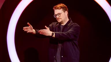 Maxi Gstettenbauer steht auf der rechten Bildseite. Er streckt beide Arme in Richtung des linken Bildrandes aus. Er ist dunkel gekleidet. Auf der rechten Seite steht ein schwarzer Barhocker, auf dem ein Mikrofon liegt. Maxi Gstettenbauer trägt eine Brille.
