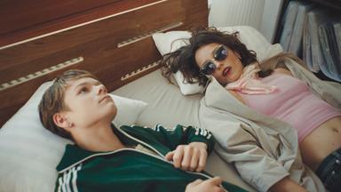 Zwei Personen liegen nebeneinander auf einem Bett mit weißen Kissen. Eine Person trägt eine grüne Trainingsjacke, die andere Sonnenbrille, Schal und helles Oberteil. Im Hintergrund sind Holzmöbel und ein Regal zu sehen.