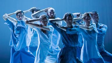 Eine Gruppe von fünf Tänzerinnen und drei Tänzern in blauen Kostümen, sie blicken gerade nach vorne, den rechten Arm angewinkelt und die Hand auf den eigenen Kopf gelegt, mit dem linken Arm den rechten Ellbogen haltend. 