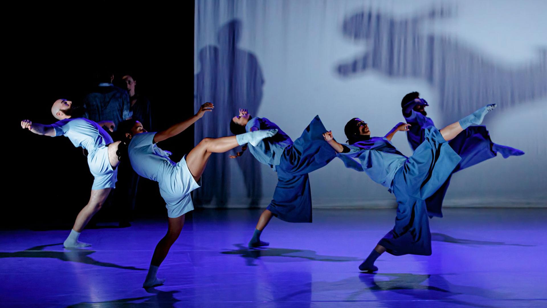 Drei Tänzerinnen und zwei Tänzer in blauen Kostümen, sie haben jeweils das rechte Bein weit angehoben und den Oberkörper zurückgebeugt. Ihre Schatten zeichnen sich auf der weißen Stoffbahn hinter ihnen ab. Hinten links im Halbdunkel ein Mann und eine Frau, der Mann mit dem Rücken zum Publikum.