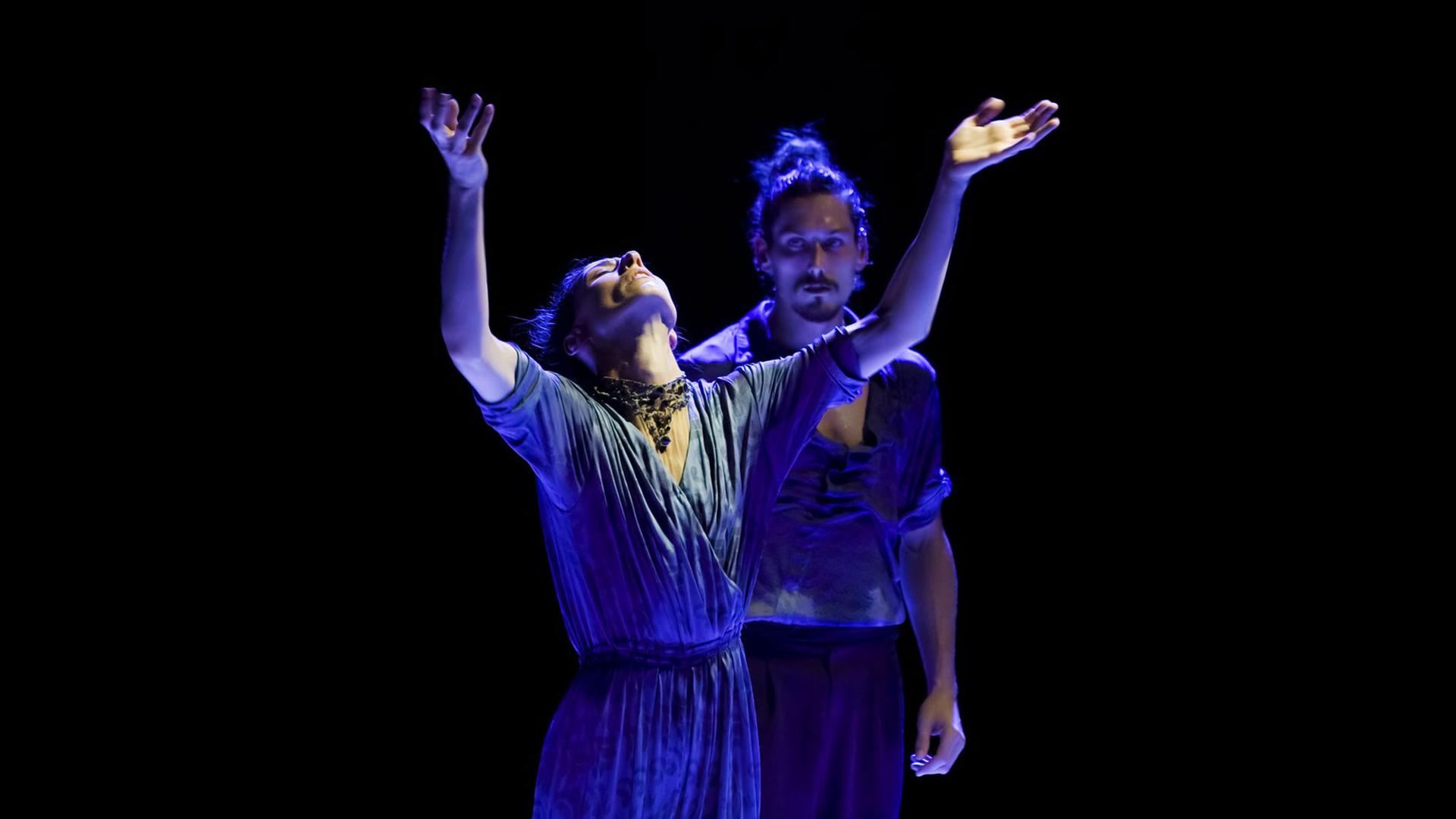 Im Vordergrund eine Frau mit erhobenen Armen und zurückgeworfenem Kopf, rechts hinter ihr ein Mann, der sie anblickt. 