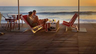 Zwei Menschen sitzen auf einer Terrasse am Meer; im Hintergrund geht die Sonne unter.
