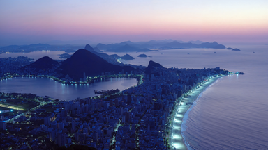 Blick auf Rio de Janeiro bei einsetzender Nacht mit beleuchteter Copacabana, Bergen im Hintergrund und dem Atlantik im Abendlicht.