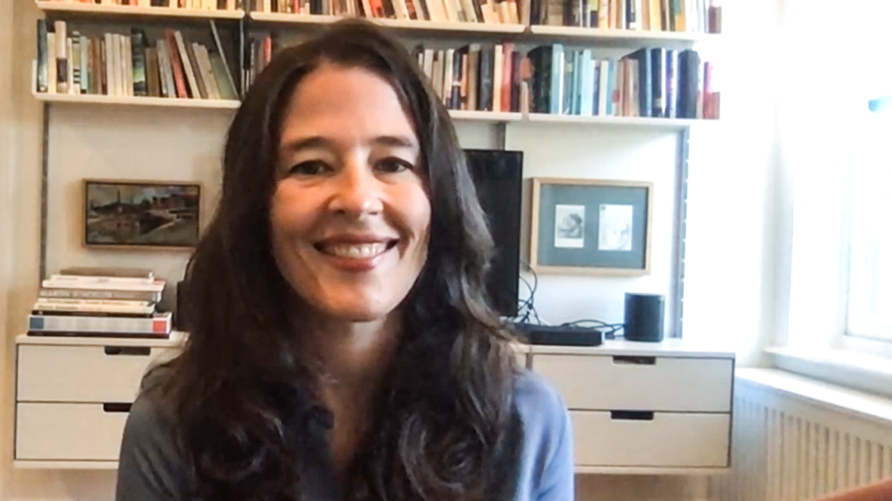 Eine lächelnde Frau mit brauem Haar sitzt vor einem Bücherregal.