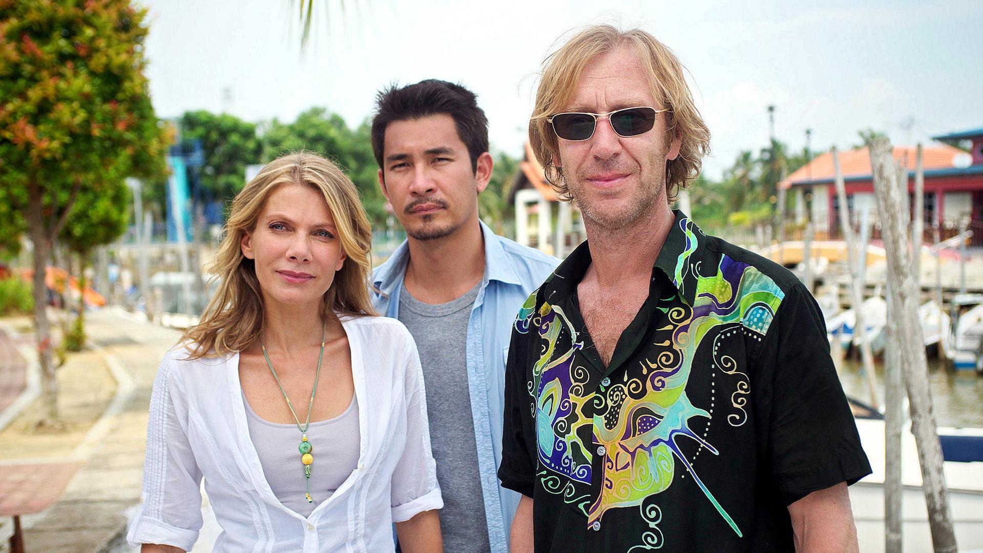 Eine blonde Frau steht mit zwei Männern an einem Boots-Steg. Einer der Männer hat asiatische Gesichtszüge, der andere trägt eine Sonnenbrille und wirkt europäisch.