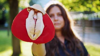 Eine Frau hält ein Stoffmodell einer Vulva in die Kamera.