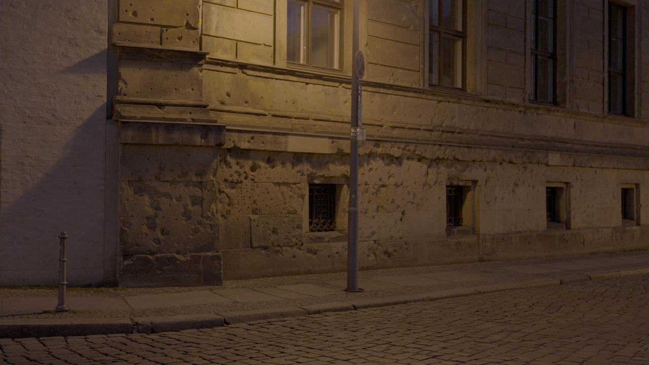Eine Hausfassade eines Altbaus in einer Berliner Straße bei Nacht, beleuchtet von Straßenlaternen. Deutlich erkennbar sind zahlreiche Einschusslöcher und Beschädigungen in der Fassade – Spuren des Zweiten Weltkriegs. Besonders im unteren Bereich der Hauswand sind die Einschläge sehr dicht, die Oberfläche ist stark zerfurcht. Vor der Fassade verläuft ein schmaler Gehweg mit Pflastersteinen, daneben eine Straße aus grobem Kopfsteinpflaster. Ein Straßenpfahl mit einem Verkehrsschild steht direkt vor der Wand, wodurch die Spuren teilweise verdeckt sind. Die Atmosphäre wirkt ruhig und beinahe still, gleichzeitig erinnern die Einschusslöcher eindrücklich an die Zerstörungen und die Gewalt der Vergangenheit.