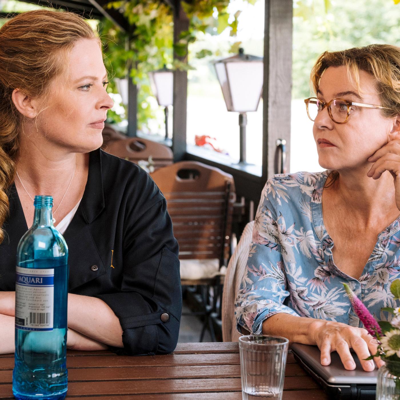 Mutter und erwachsene Tochter sitzen auf der Terrasse eines Cafés und schauen sich fragend an