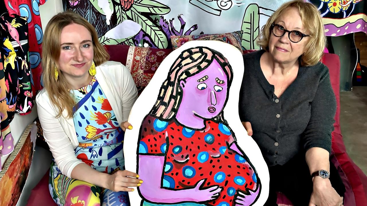 Zwei Frauen sitzen nebeneinander auf einem Sofa und halten gemeinsam ein farbenfrohes Gemälde einer lila Frau mit rotem Kleid; der Hintergrund ist mit bunten Mustern dekoriert.