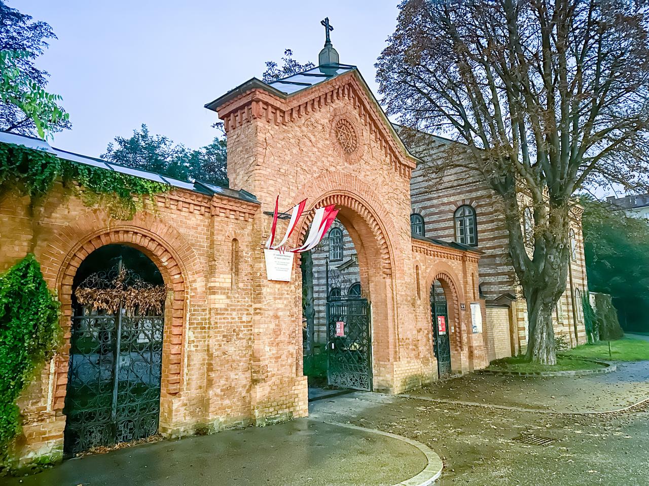 Das Bild zeigt das Eingangstor des Friedhofs St. Marx in Wien. Das Tor besteht aus roten Backsteinen und ist mit einem geschwungenen Bogen versehen. Es gibt dekorative Elemente, darunter ein Kreuz an der oberen Mitte des Tors. An den Seiten des Eingangs sind Metallgittertüren sichtbar, hinter denen sich die Sicht auf den Friedhof verbirgt.   Über dem Tor hängt eine Tafel, die vermutlich Informationen zum Friedhof enthält. Am Tor sind außerdem einige Flaggen angebracht, die sich sanft im Wind bewegen. Im Hintergrund ist ein weiteres Gebäude zu sehen, das ebenfalls aus Backstein besteht und große Fenster hat. Es ist von Bäumen umgeben, die erste herbstliche Blätter verloren haben. Der gesamte Eindruck ist von einer ruhigen und nachdenklichen Atmosphäre geprägt, passend zum Thema des Films, der sich mit Tod und Erinnerung in der Musik beschäftigt. Die Lichtverhältnisse sind gedämpft, was die melancholische Stimmung unterstreicht.