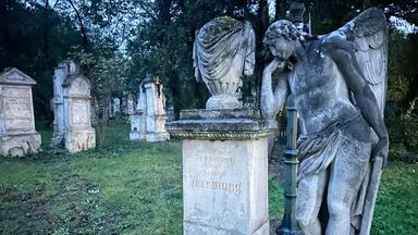 Das Bild zeigt eine Szene auf einem Friedhof, wahrscheinlich am Friedhof St. Marx. Im Vordergrund steht eine detaillierte Skulptur eines Engels, der nachdenklich mit einer Hand an seinem Kopf lehnt. Die Statue ist aus Stein und wirkt verziert und alt. Neben dem Engel befindet sich ein monumentaler Grabstein mit der Inschrift „Auferstehung“ in goldener Schrift.   Im Hintergrund sind mehrere Grabsteine zu sehen, die unterschiedlich geformt und angeordnet sind, und die Grabinschriften sind teilweise nicht klar lesbar. Die Umgebung ist grün, mit Gras und einigen Bäumen, die eine ruhige und friedliche Atmosphäre schaffen. Es scheint Dämmerung zu sein, was dem Bild eine melancholische Stimmung verleiht.   Die Gesamtszene vermittelt ein Gefühl von Trauer, aber auch von Trost durch die künstlerische Darstellung des Engels und den historischen Kontext des Ortes.