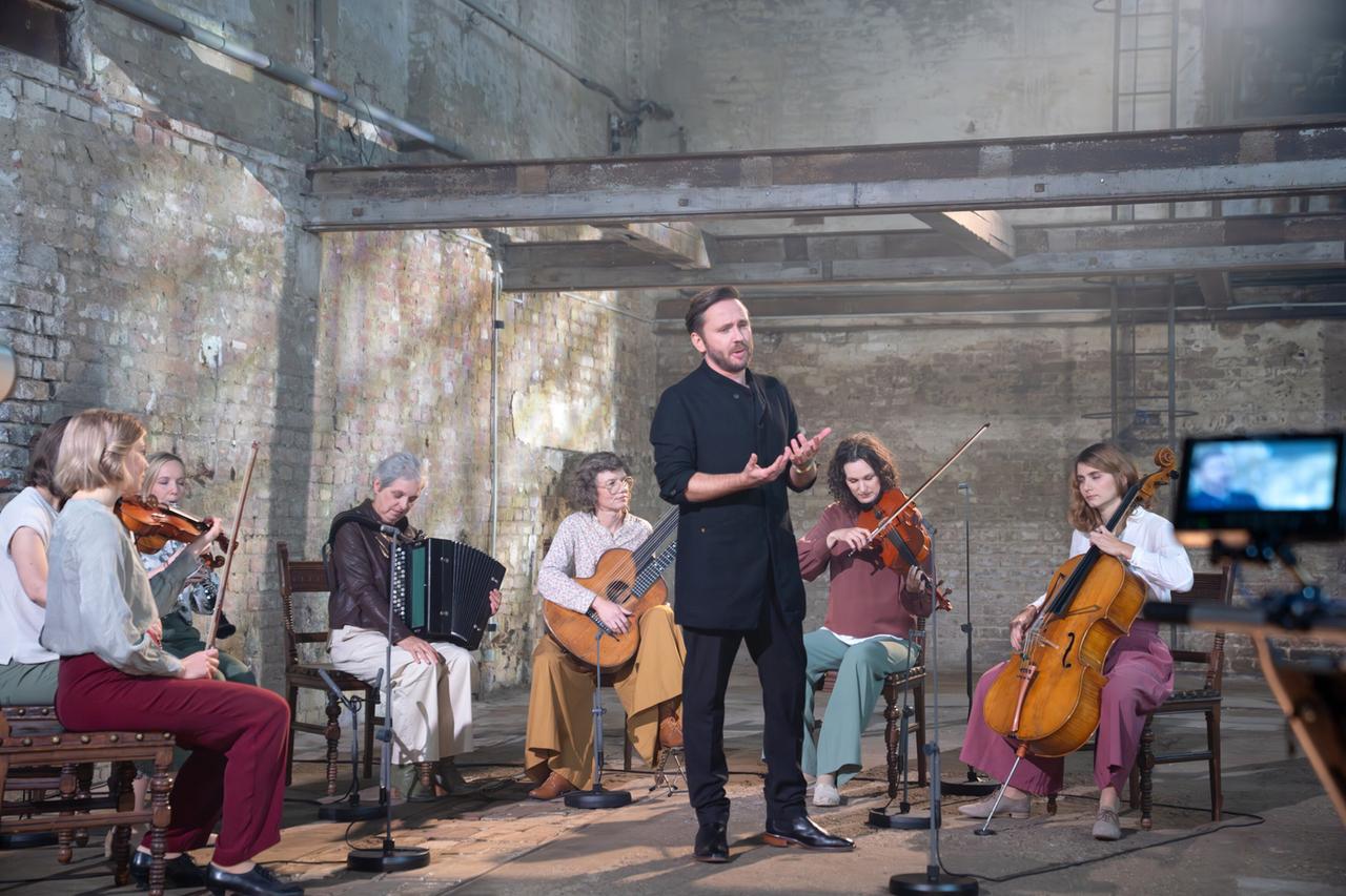 Das Bild zeigt eine musikalische Darbietung in einem offenen, lichtdurchfluteten Raum mit einer rauen Ziegelsteinwand. In der Mitte des Bildes steht ein Mann in einem schwarzen Outfit, der anscheinend spricht oder singt. Um ihn herum sitzen fünf Musikerinnen.   Von links nach rechts sind das folgende Instrumente zu sehen: - Eine Frau spielt Geige. - Eine weitere Frau hat eine zweite Geige in der Hand. - Eine dritte Musikerin spielt ein Akkordeon. - Eine Frau mit einer Gitarre sitzt daneben. - Die letzte Musikerin spielt ein Cello.  Sie sind auf Stühlen platziert, während Mikrofone um sie herum aufgestellt sind. Der Boden des Raumes ist aus Holz und der gesamte Raum wirkt aufgrund der Wände und Lichtverhältnisse eher industriell. Es sind keine speziellen Identifikationen der Musiker oder Informationen zu ihrem Aussehen zu sehen, die spezifische Merkmalen beschreiben.
