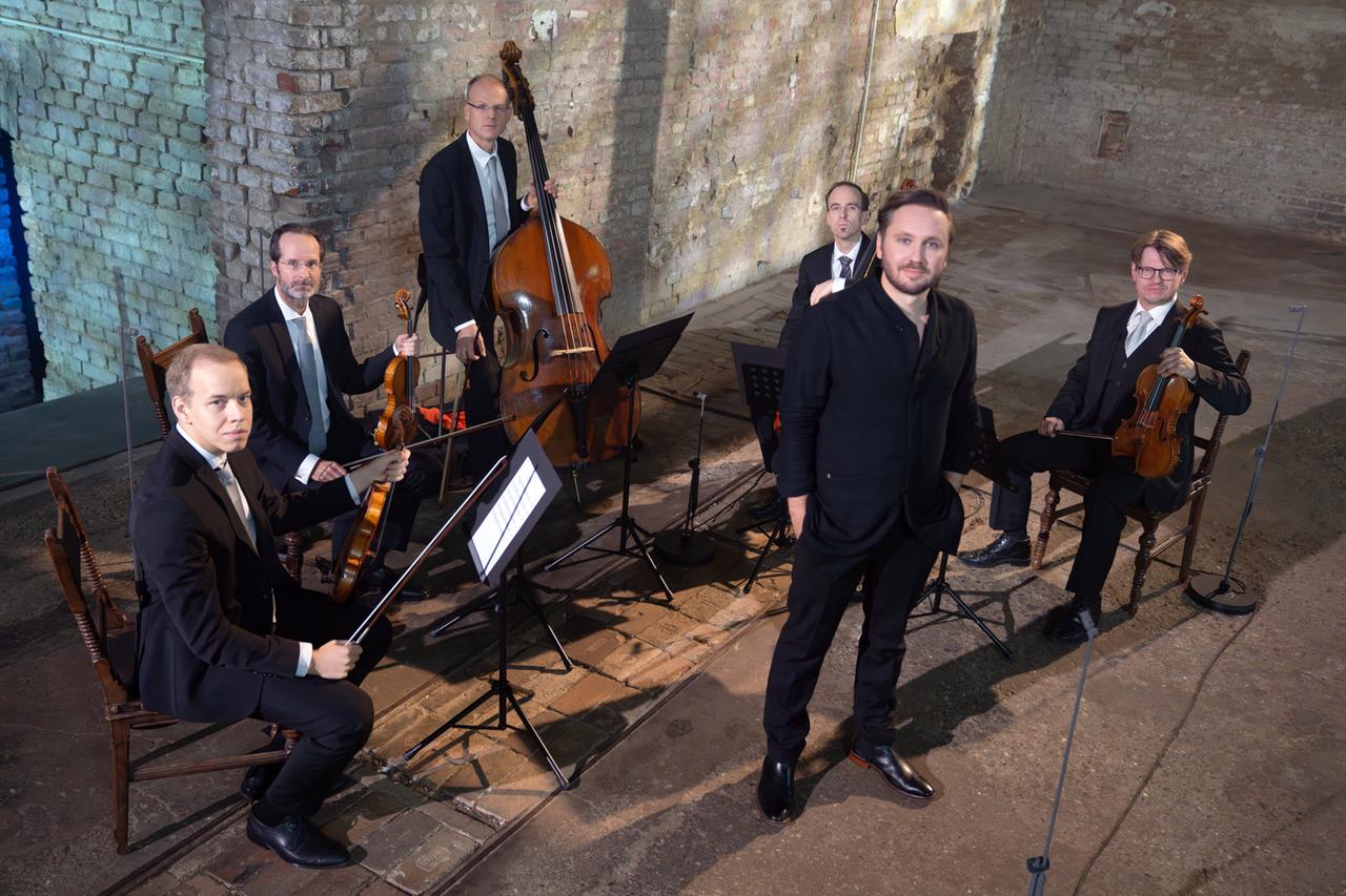 Das Bild zeigt eine Gruppe von sechs Musikern in formeller Kleidung, die in einem historischen Raum mit Ziegelwänden stehen und sitzen. Im Vordergrund befindet sich ein Mann, der ein schwarzes Outfit trägt und in einer entspannten Pose steht. Hinter ihm sind fünf weitere Musiker zu sehen, die in einem Halbkreis angeordnet sind.   Zwei Musiker halten Streichinstrumente: einer mit einer Violine und ein anderer mit einem Cello. Die restlichen Musiker sind an Stühlen platziert und haben verschiedene Instrumente oder Notenständer vor sich. Der Raum hat einen rustikalen Charakter mit einem steinernen Boden und ist gut beleuchtet, wobei Licht von einer Seite auf die Szene fällt.   Hintergrunddetails zeigen die Ziegelwand des Raumes, die etwas unregelmäßig und verwittert aussieht. Es sind keine spezifischen Merkmale oder Dekorationen im Hintergrund zu erkennen, die auf einen bestimmten Ort hindeuten. Der Gesamteindruck vermittelt eine Atmosphäre der Musikalität und des Zusammenkommens.