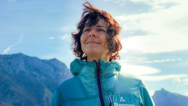Das Bild zeigt eine Frau, die im Freien vor einer beeindruckenden Berglandschaft steht. Sie hat lockiges, braunes Haar, das vom Wind aufgewirbelt wird. Die Frau trägt eine grüne Jacke, die warm und funktional aussieht, und lächelt leicht, während sie in die Ferne blickt. Im Hintergrund sind große, felsige Berge zu sehen, die teilweise mit Schnee bedeckt sind. Der Himmel ist teilweise bewölkt mit einigen kleinen, weißen Wolken und hat eine insgesamt helle, freundliche Atmosphäre. Die Umgebung wirkt natürlich und unberührt, was auf eine alpine Landschaft hindeutet.