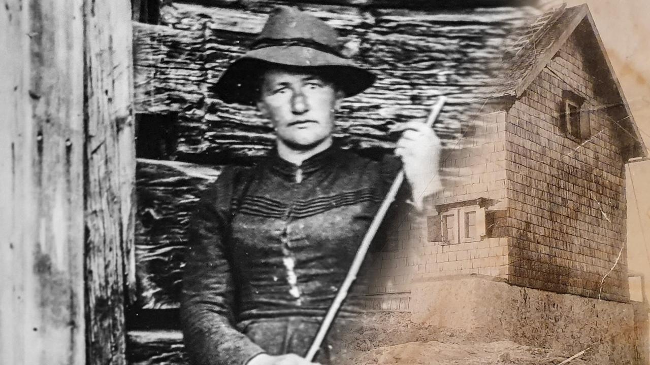 Das Bild zeigt eine historische schwarz-weiße Fotografie von einer Frau, die in traditioneller Kleidung abgebildet ist. Sie steht vor einer hölzernen Wand, die aus grob behauenen Brettern besteht. Die Frau trägt einen breitrandigen Hut und hat eine ernste Miene. In der rechten Hand hält sie einen langen Stock oder Besen. 

Im Hintergrund ist ein charakteristisches, verputztes Gebäude zu sehen, das sich von der Holzstruktur abhebt und mit schindelartigen Materialien verkleidet ist. Das Gebäude hat Fenster mit leicht hervorstehenden Fensterläden und steht auf einem stufenartigen Fundament. 

Die gesamte Szene vermittelt einen historischen Eindruck und hat einen Bezug zur Bergsteiger-Geschichte Österreichts, insbesondere zu den Leistungen von Frauen in diesem Bereich.