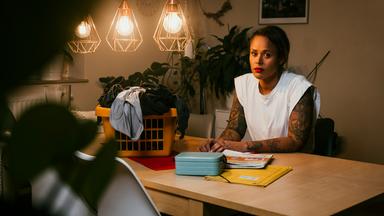 Tamika Förster sitzt abends bei künstlichem Licht an einem Esstisch. Vor ihr auf dem Tisch liegen Arbeitsmaterialien, zwei Kinderbrotdosen, daneben steht der Wäschekorb. Tamika sinniert.