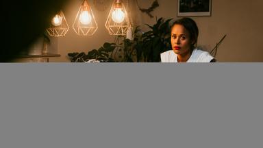 Tamika Förster sitzt abends bei künstlichem Licht an einem Esstisch. Vor ihr auf dem Tisch liegen Arbeitsmaterialien, zwei Kinderbrotdosen, daneben steht der Wäschekorb. Tamika sinniert.