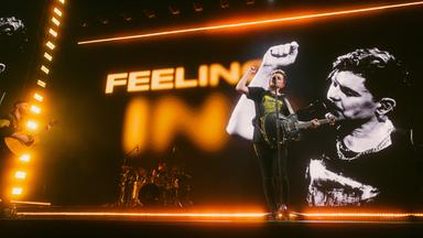 Der Sänger Michael Patrick Kelly steht mit einer Gitarre auf der Bühne, singt und hält die rechte Faust in die Luft.