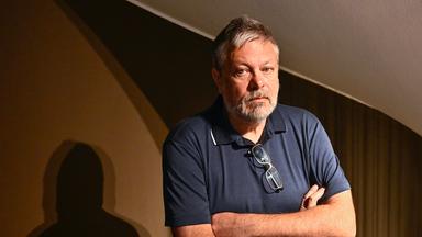 Foto eines Mannes mit graumeliertem Bart und Haaren, der mit verschränkten Armen dasteht und ernst in die Kamera schaut.