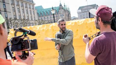 Ein Mann steht vor einer gelben Kunstinstallation und spricht in ein Mikrofon, während er von zwei Kameras gefilmt wird.