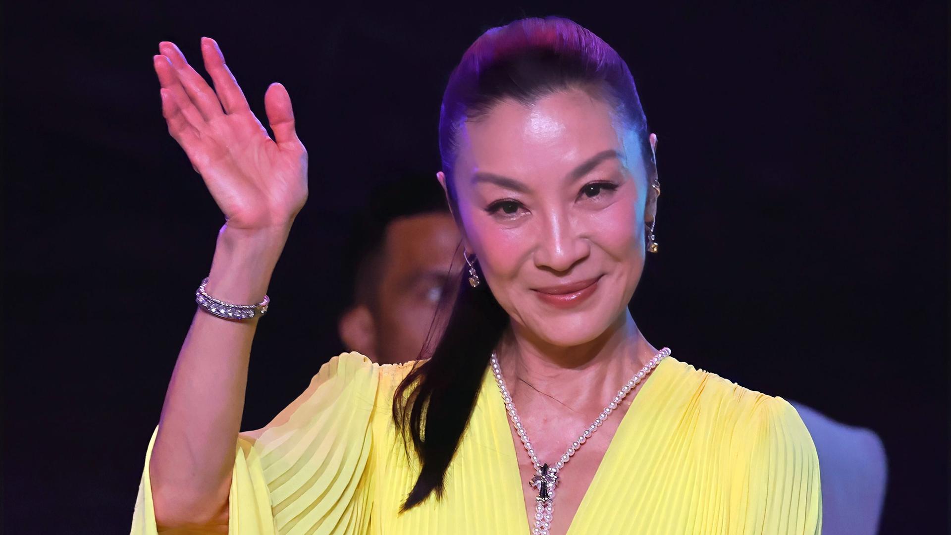 Eine asiatische Frau mit langem Pferdeschwanz trägt ein schickes gelbes Kleid und Perlenschmuck und winkt lächelnd mit der rechten Hand.