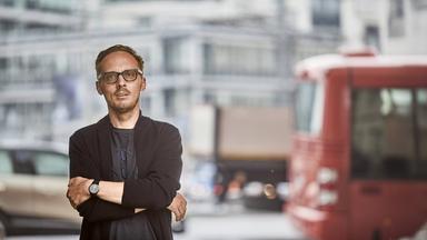 Ein Mann mit Brille und Dreitagebart, leger gekleidet mit einem schwarzen T-Shirt und schwarzem Sakko steht mit verschränkten Armen vor einem Fenster, hinter dem verschwommen eine Straße in der Stadt zu sehen ist.