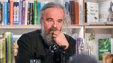 Ein Mann mit graumeliertem Bart und Haaren sitzt an einem Tisch mit Mikrofon und fasst sich nachdenklich mit einer Hand ans Kinn. Hinter ihm befindet sich ein Bücherregal.