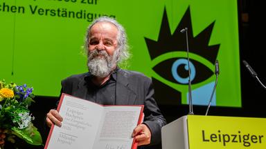 Ein Mann mit graumeliertem Bart und Haaren präsentiert auf einer Bühen eine Kladde mit einer Urkunde. Hinter ihm ist ein grünes Plakat der Leipziger Buchmesse zu sehen.