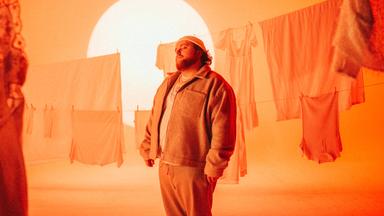 Ein korpulenter Mann in Jogginghose und weiter Jacke steht mit geschlossenen Augen verträumt vor einer Wäscheleine. Hinter ihm brennt die Sonne und taucht alles in ein orange-gelbes Licht