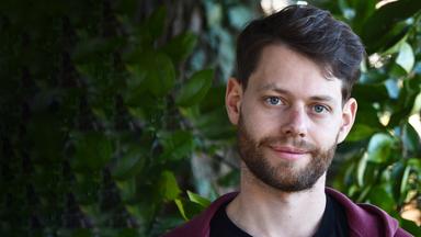 Ein junger Mann mit Bart und braunem Haar steht vor einem Hintergrund aus grünen Pflanzen und blickt mit lächelnd in die Kamera.