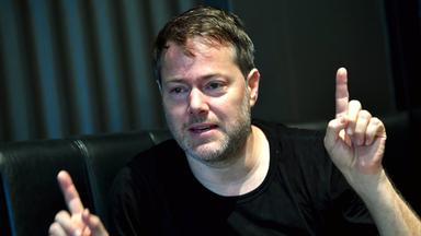 Ein Mann mit schwarzem T-Shirt spricht und gestikuliert mit seinen Händen.