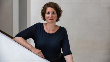 Eine Frau mit kinnlangen dunkelbraunen Haaren und rotem Lippenstift lehnt sich an das weiße Geländer einer Treppe.