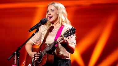 Miss Allie steht mit Gitarre in der Mitte des Bildes. Vor ihr ist ein Mikrofon. Der Hintergrund ist rot beleuchtet.