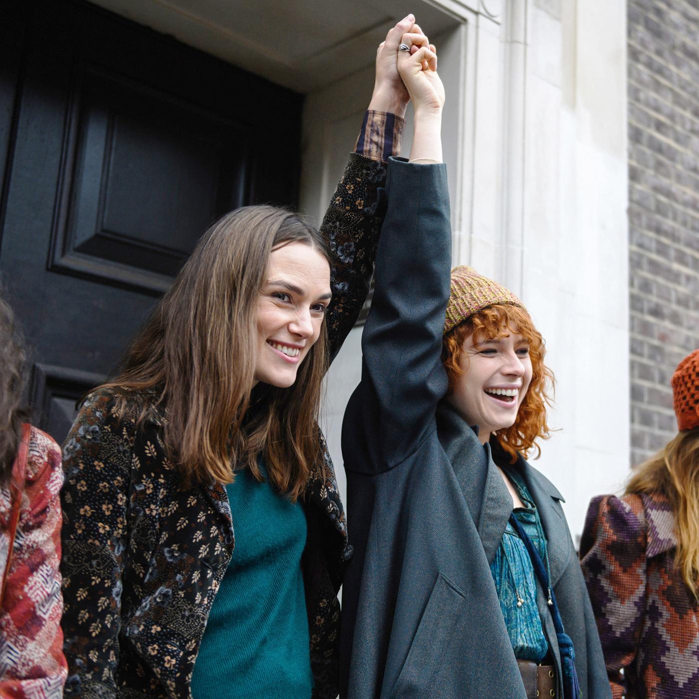 Frauen vor einem alten Gebäude. Die zwei mittleren Frauen halten sich an den Händen und heben die verbundenen Arme nach oben.