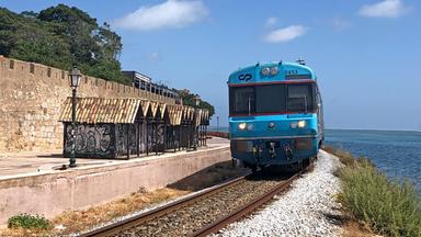 Ein Zug auf Schienen, auf der einen Seite das Meer, auf der anderen Seite bunt besprühte kleine Hütten und eine hohe Mauer