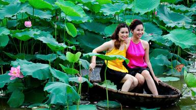 Zwei junge Frauen in gelben und pinken Tops sitzen in einem kleinen Boot uns lassen sich inmitten von Blättern und Blumen im Westlake am Stadtrand von Hanoi fotografieren.