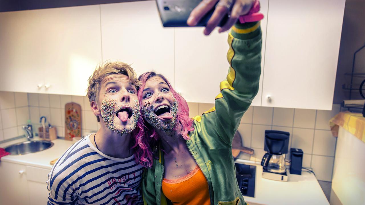 Das Bild zeigt zwei jugendliche Personen in einer modernen, hellen Küche. Die Wände sind in einem neutralen Farbton gehalten, und es gibt weiße Küchenschränke sowie eine Spüle und Kochstelle im Hintergrund.   Die männliche Person, die rechts steht, hat blondes, leicht gewelltes Haar und trägt ein gestreiftes T-Shirt. Der Gesichtsausdruck ist lächelnd und jugendlich ausgelassen. Die weibliche Person, die links steht, hat kräftig gefärbte, pinke Haare und trägt einen grünen Kapuzenpullover über einem orangenen Oberteil. Beide haben sichtbare bunte Streusel auf ihren Gesichtern verteilt, als ob sie gerade ein Spiel oder eine lustige Aktivität gemacht hätten. Sie ziehen die Zunge heraus und lachen, während sie ein Selfie machen.  Das Bild vermittelt eine fröhliche und unbeschwerte Atmosphäre, die die Leichtigkeit des Lebens in der Jugend widerspiegelt.