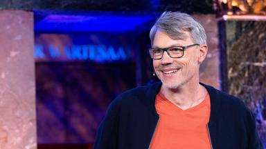 Gastgeber Christoph Sieber in dunkler Strickjacke und orangefarbenem T-Shirt steht lächelnd im Studio.