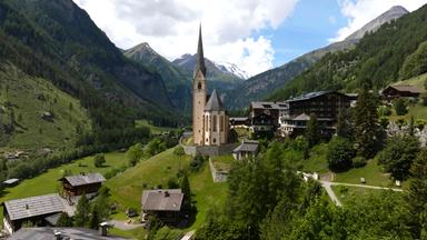 Das Bild zeigt eine malerische alpine Landschaft mit einer charakteristischen Pfarrkirche im Vordergrund. Die Kirche hat einen hohen, spitzen Turm und ist aus hellen Steinen erbaut, mit einigen dunklen Dachelementen. Im Hintergrund sind dramatische Berggipfel zu sehen, die teilweise schneebedeckt sind. Die Landschaft um die Kirche ist grün und bewaldet, mit sanften Hügeln und vereinzelten Häusern im Vordergrund. Diese Hütten sind traditionell aus Holz gebaut und haben Steildächer. Der Himmel ist teilweise bewölkt, was dem Bild eine dynamische Lichtstimmung verleiht. Diese Szene vermittelt ein Gefühl von Ruhe und Naturnähe in den Alpen.