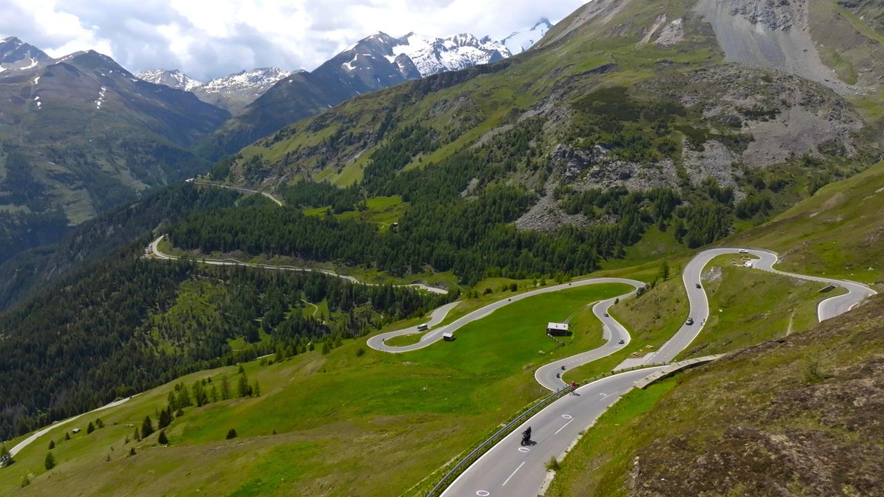Das Bild zeigt eine kurvenreiche Bergstraße, die durch eine grüne, alpine Landschaft führt. Die Straße schlängelt sich über sanfte Hügel und ist von Wiesen und vereinzelten Bäumen umgeben. Im Hintergrund erheben sich beeindruckende, schneebedeckte Berge unter einem teilweise bewölkten Himmel. Es sind mehrere Fahrzeuge sichtbar, darunter Autos und Motorräder, die die Straße befahren. Ein kleines, weißes Gebäude steht am Straßenrand, umgeben von saftigem Grün. Die gesamte Szene vermittelt das Gefühl von unberührter Natur und lädt zu Aktivitäten in der freien Natur ein.