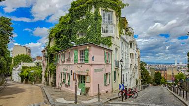 Eine kopfsteingepflasterte Gasse in Montmartre mit einem rosa Café an einer Straßenecke. Efeu bedeckt die darüberliegenden Fassaden, im Hintergrund öffnet sich der Blick über Paris.