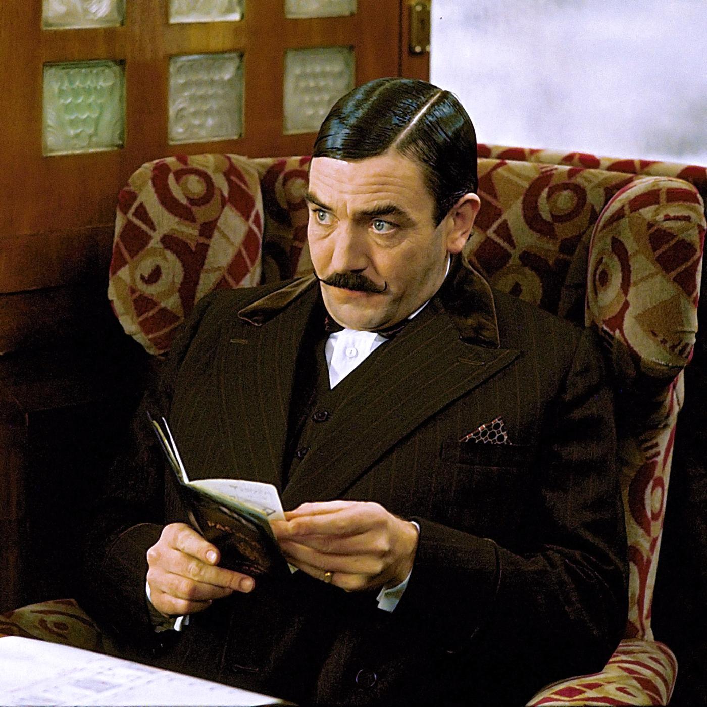 Ein Mann mit Schnurrbart und dunklem Anzug sitzt skeptisch blickend in einem Sessel an einem Tisch, einen Bleistift in der Hand.