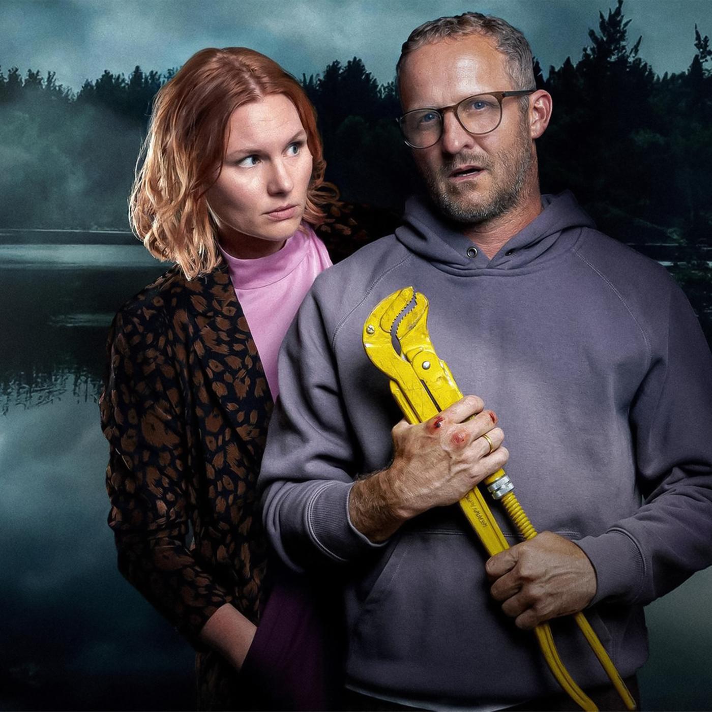 Eine Frau und ein Mann vor einem dunklen See im Wald. Der Mann hält ein großes gelbes Werkzeug in den Händen.