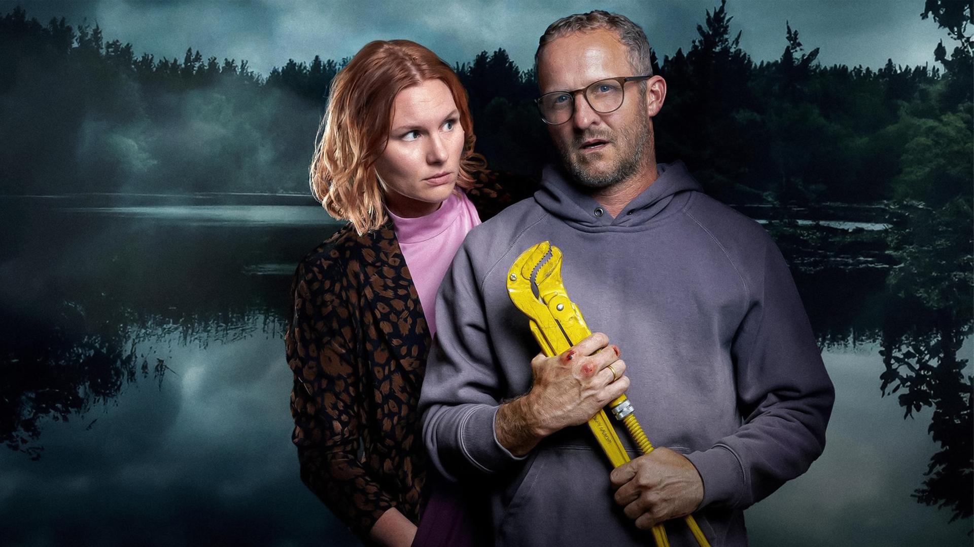 Eine Frau und ein Mann vor einem dunklen See im Wald. Der Mann hält ein großes gelbes Werkzeug in den Händen.
